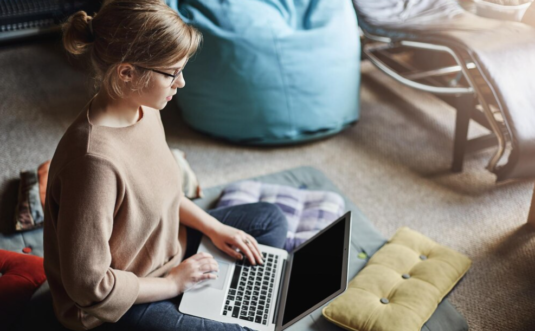woman in the cozy outfit and glasses sitting on the floor with a laptop on lap
