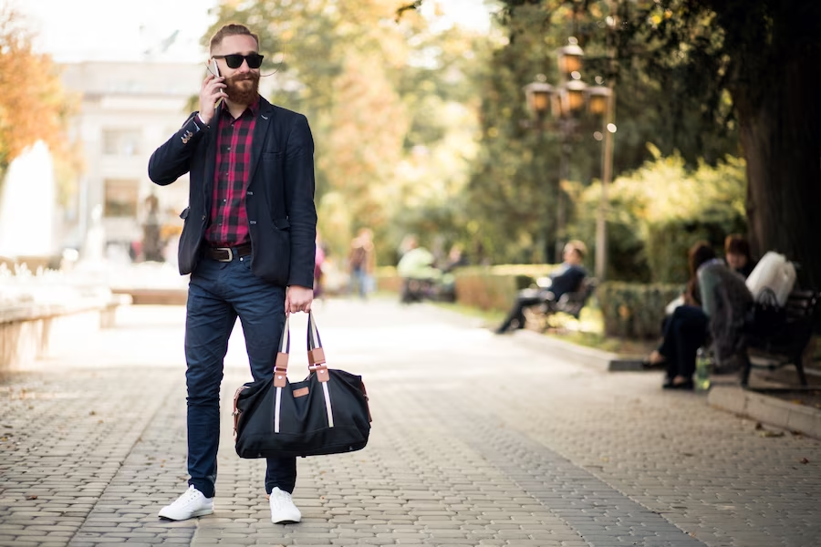 A man wearing a coat with a checkered polo underneath, standing in a park walk, holding a bag and talking on a phone