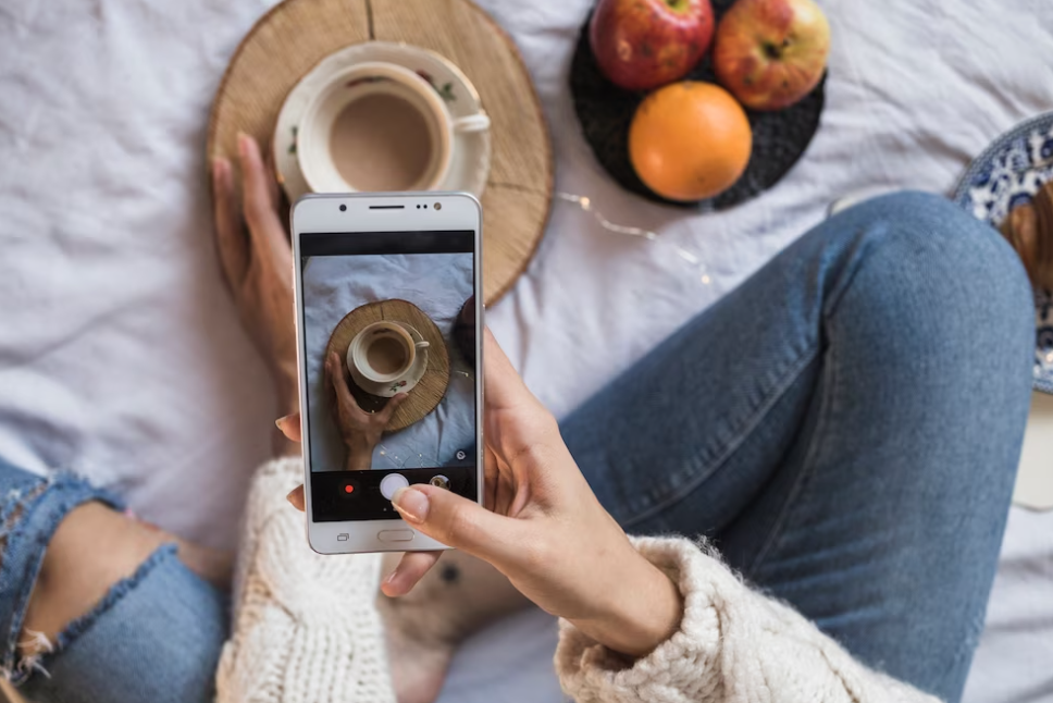 woman holds a phone and taking photo of a cup of coffee, a plate with apples on the bed