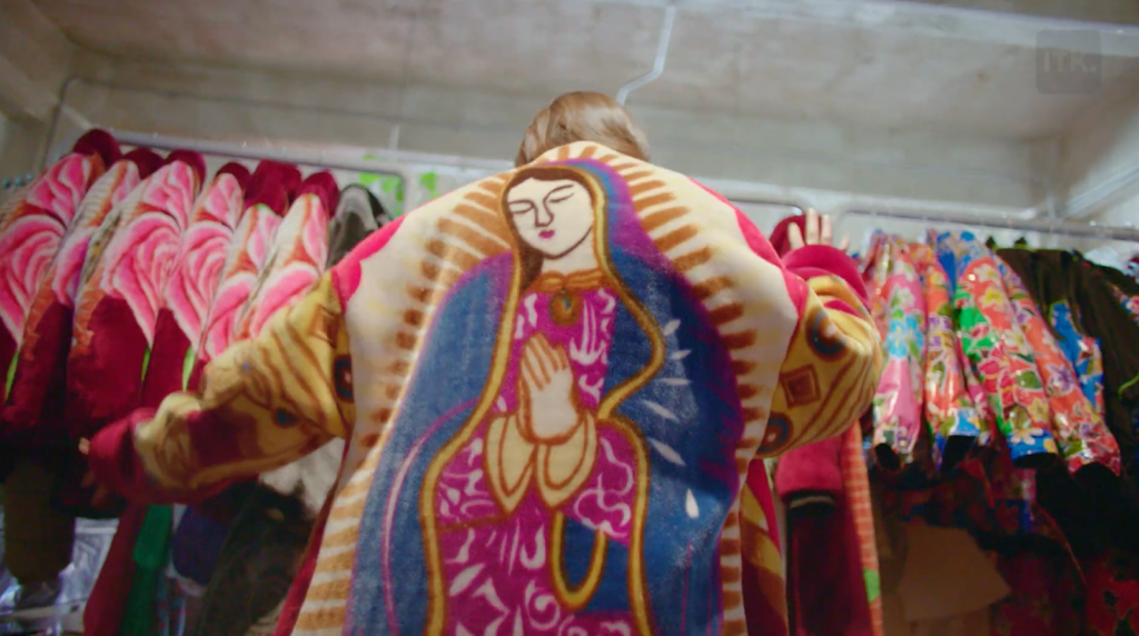 woman dancing in a colorful coat with an icon of the saint on it, coats on hangers behind