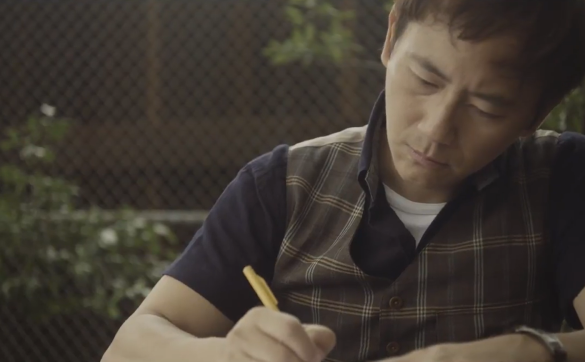 kai d in a waistcoat and blue t-shirt  sitting outdoors and making notes