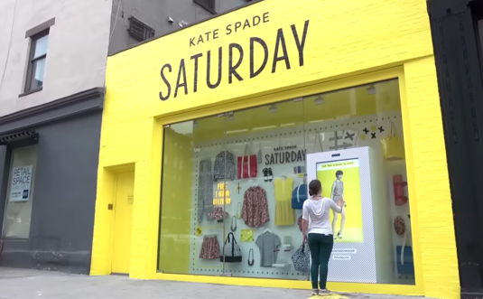 woman standing on the street near the yellow building with Kate Spade Saturday words on it